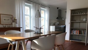 A modern dining area with a wooden table and white chairs, a kitchen in the background, and a bookshelf with books. - Vacation home 3 - Holiday homes Michaelis 1 to 4 in Quilitz (Rankwitz)