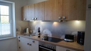 A kitchen with wooden cabinets, a built-in oven, a coffee maker, and a wine bottle. - Haus an der Pferdekoppel (Fehmarn)