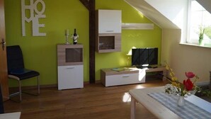 A room with a green wall, wooden beams, a TV, a desk, a chair, and a window with a plant. - Haus an der Pferdekoppel (Fehmarn)