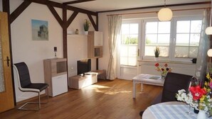 A room with wooden flooring, a white table with a blue checkered tablecloth, a black chair, a white cabinet, and a window with curtains. - Haus an der Pferdekoppel (Fehmarn)