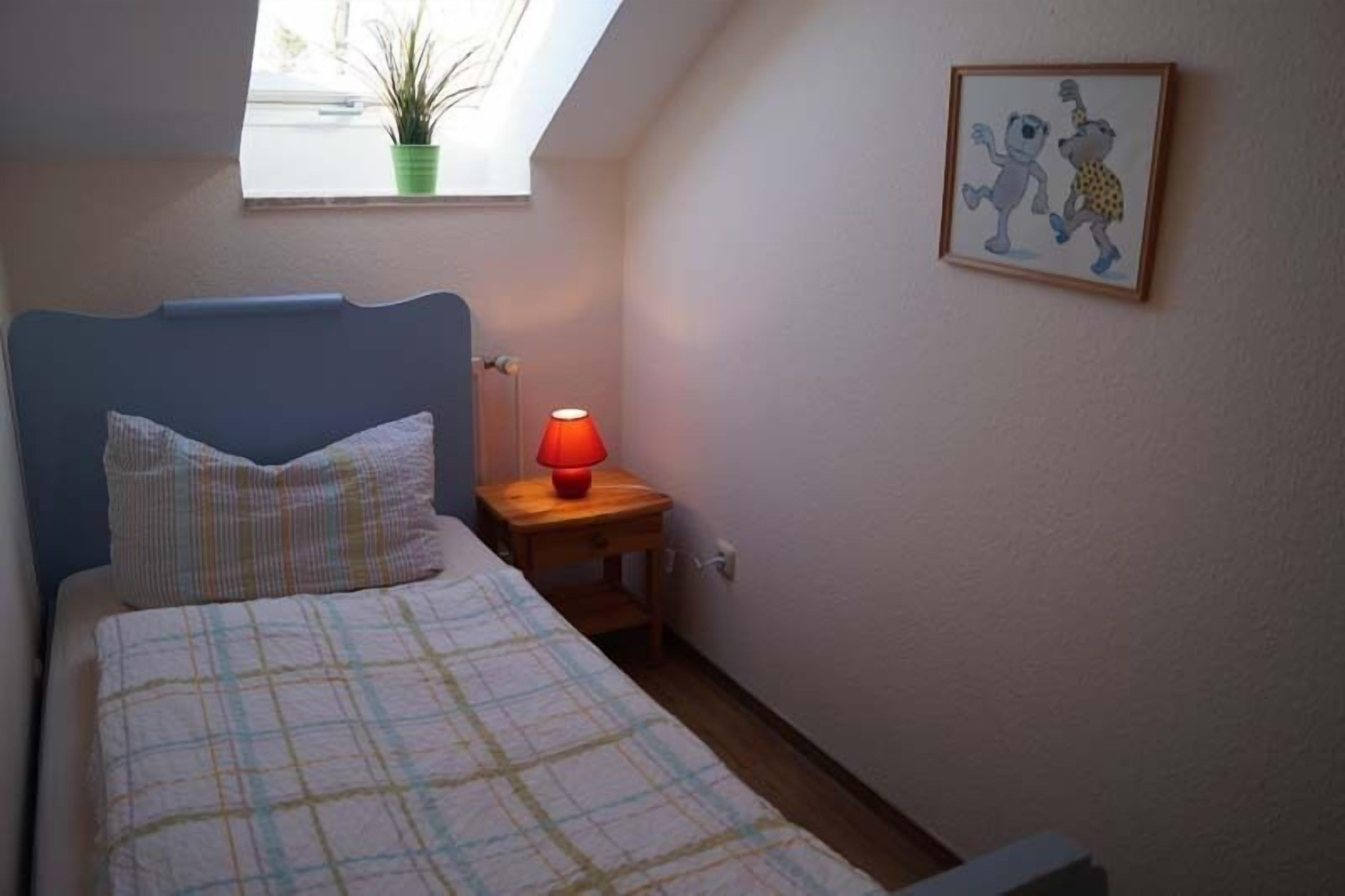 Ett sovrum med en säng, ett nattduksbord med en lampa, ett fönster med en växt och en inramad tavla på väggen.
