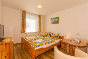 Un dormitorio con cama de madera, una mesita redonda pequeña, una silla, un televisor y una ventana con cortinas.