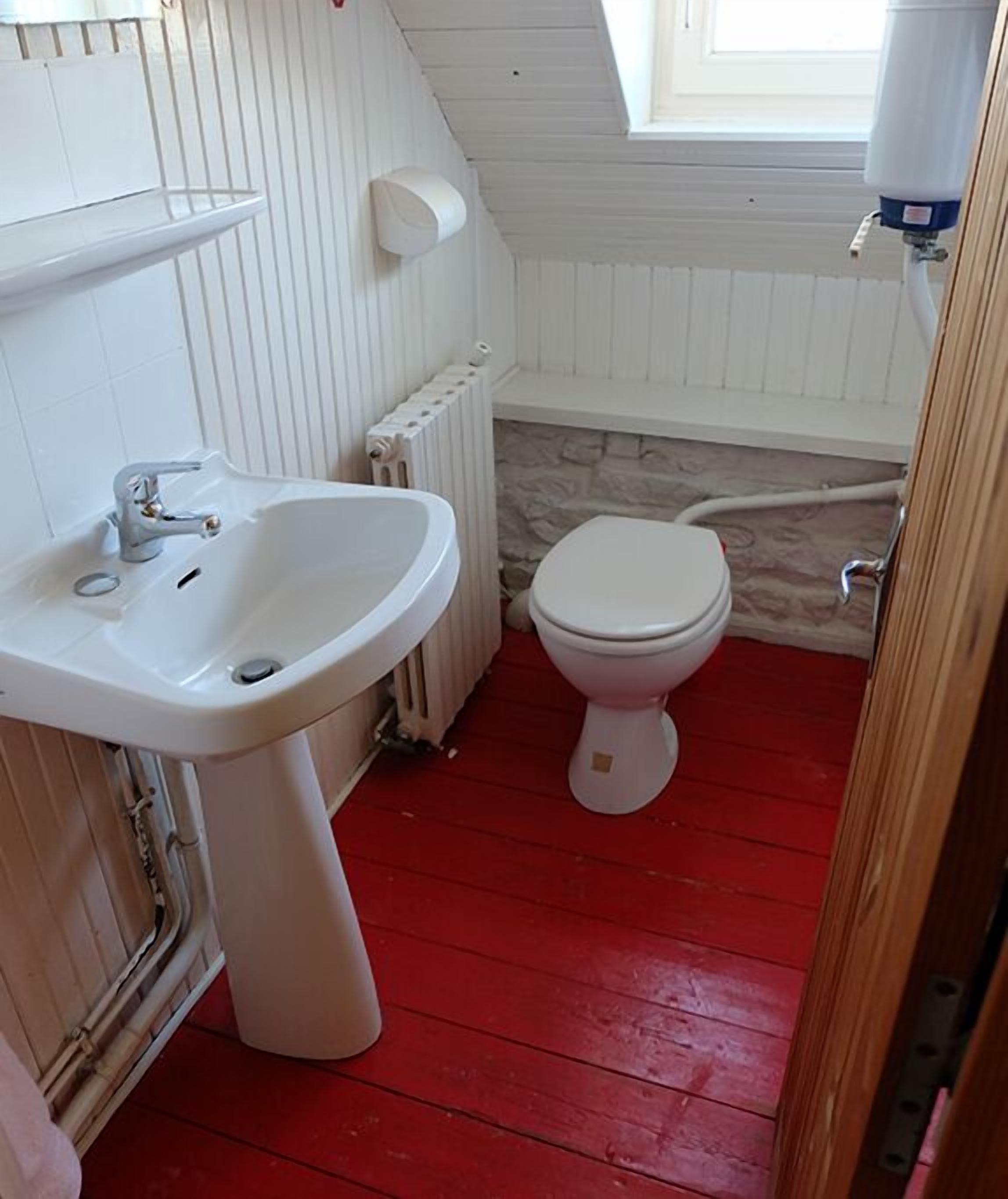 A small, compact bathroom with a white sink, toilet, and radiator. The floor is red, and there is a skylight above the toilet.