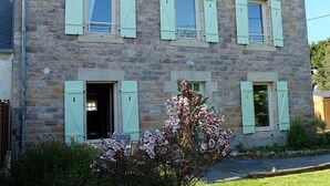 A two-story stone building with light-colored shutters, a visible entrance, and a small garden with flowering plants. - The Hauts de Kerity, 1920's house by the sea (Paimpol)