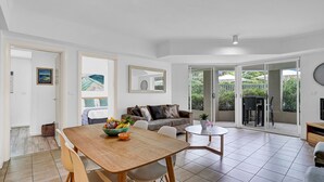 A spacious living area with a wooden dining table, a sofa, and a small round table. There is a balcony with a view of greenery.