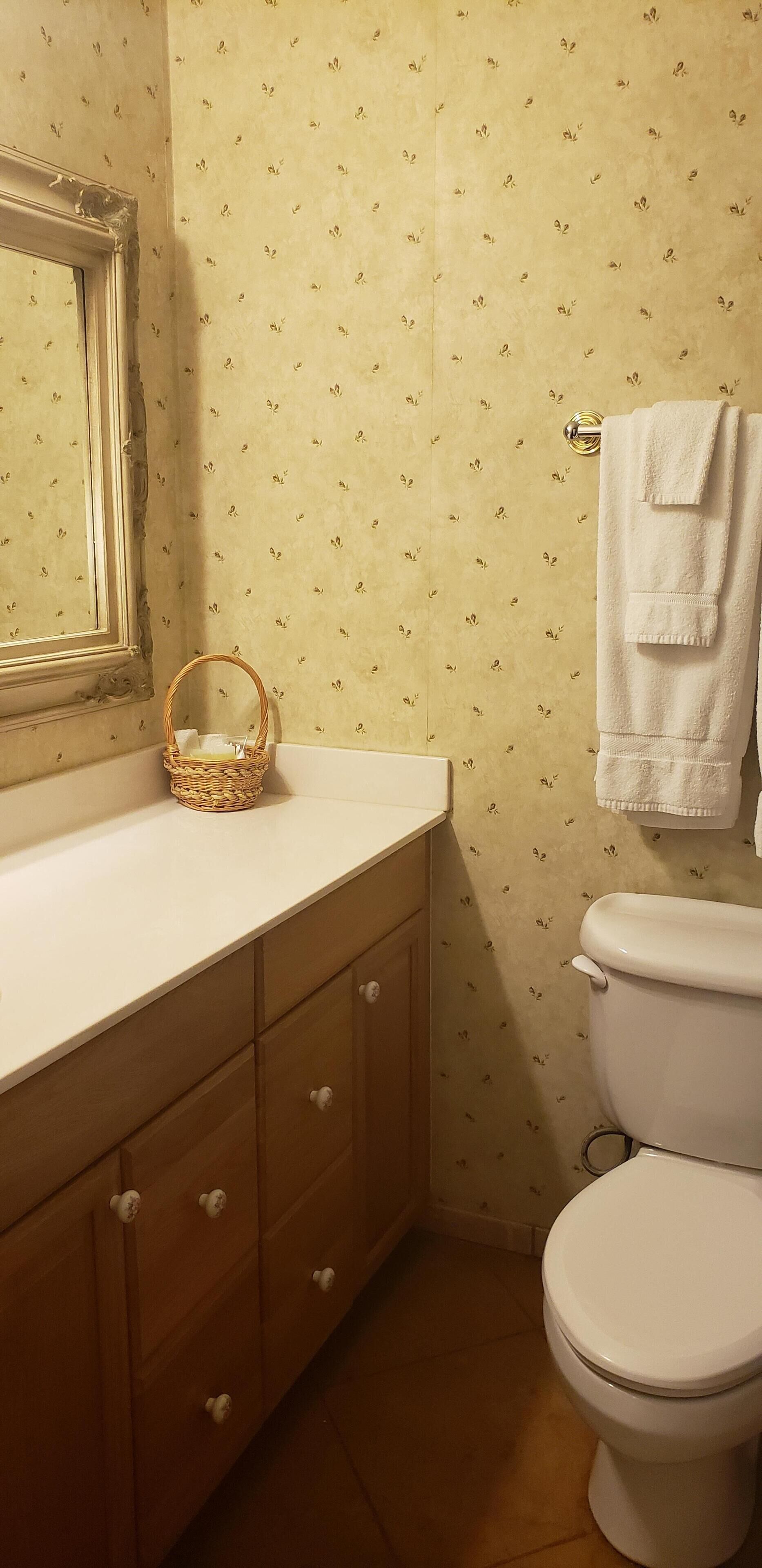 Une salle de bain avec des toilettes blanches, un lavabo avec un plan de toilette blanc, un meuble en bois avec des tiroirs, un panier en osier posé sur le plan de toilette et un mur orné d’un motif floral.