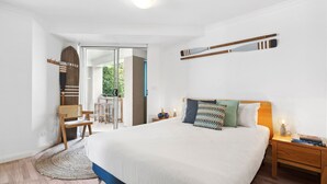 Une chambre spacieuse avec un grand lit, une tête de lit en bois, une table de chevet avec une lampe et une vue sur l’extérieur grâce à une porte vitrée.