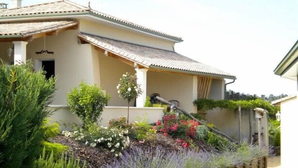 Une maison avec un toit en tuiles, un porche et un jardin où poussent différentes plantes et fleurs.