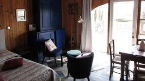 Een knus interieur met houten wanden, een blauwe kast, een bed met beddengoed met een patroon, een kleine tafel met stoelen, en uitzicht op het buitenlands gebied door een deur.