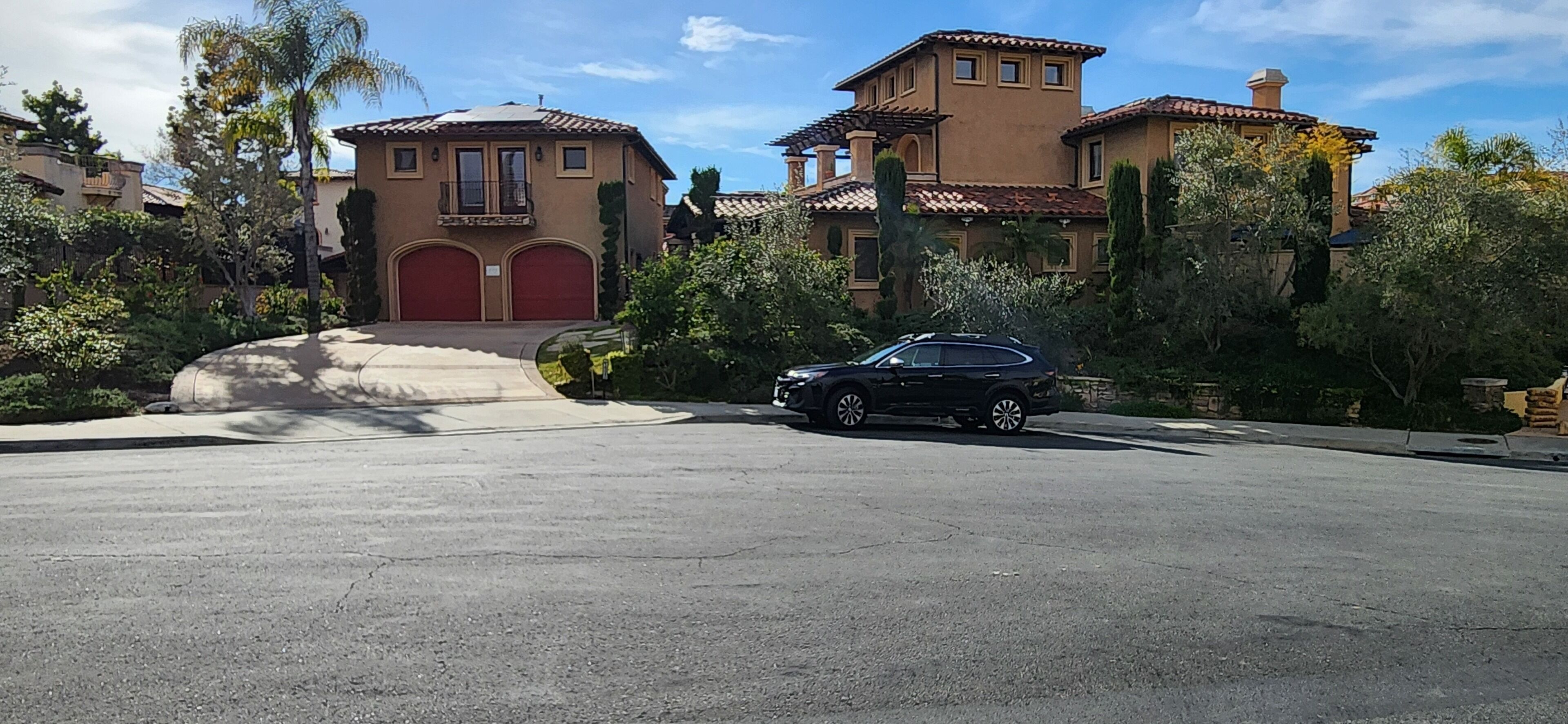 A large, two-story house with a red garage door, surrounded by lush greenery and a clear blue sky.