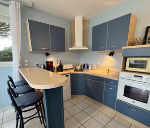 A modern kitchen with blue cabinets, a central island with bar stools, a built-in oven, and a microwave. - HOUSE NEAR THE SEA (Portbail)
