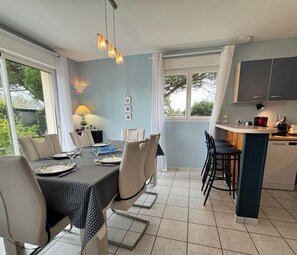 A modern dining area with a table set for four, a kitchen with a bar area, and large windows offering a view of the outdoors. - HOUSE NEAR THE SEA (Portbail)