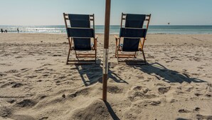 Una playa con dos sillas de madera bajo un paraguas oscuro, arena y el océano al fondo.