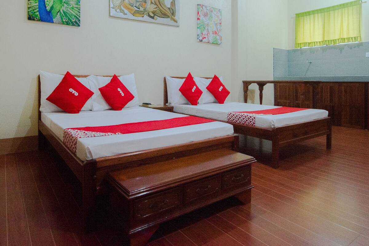A hotel room with two wooden beds, each with red pillows and a red bedspread. There are two paintings on the wall, a wooden bench, and a window with a yellow curtain.