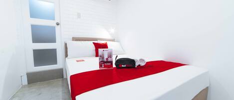 A hotel room with a bed, a red blanket, a pillow, a bag, and a box on the bed.