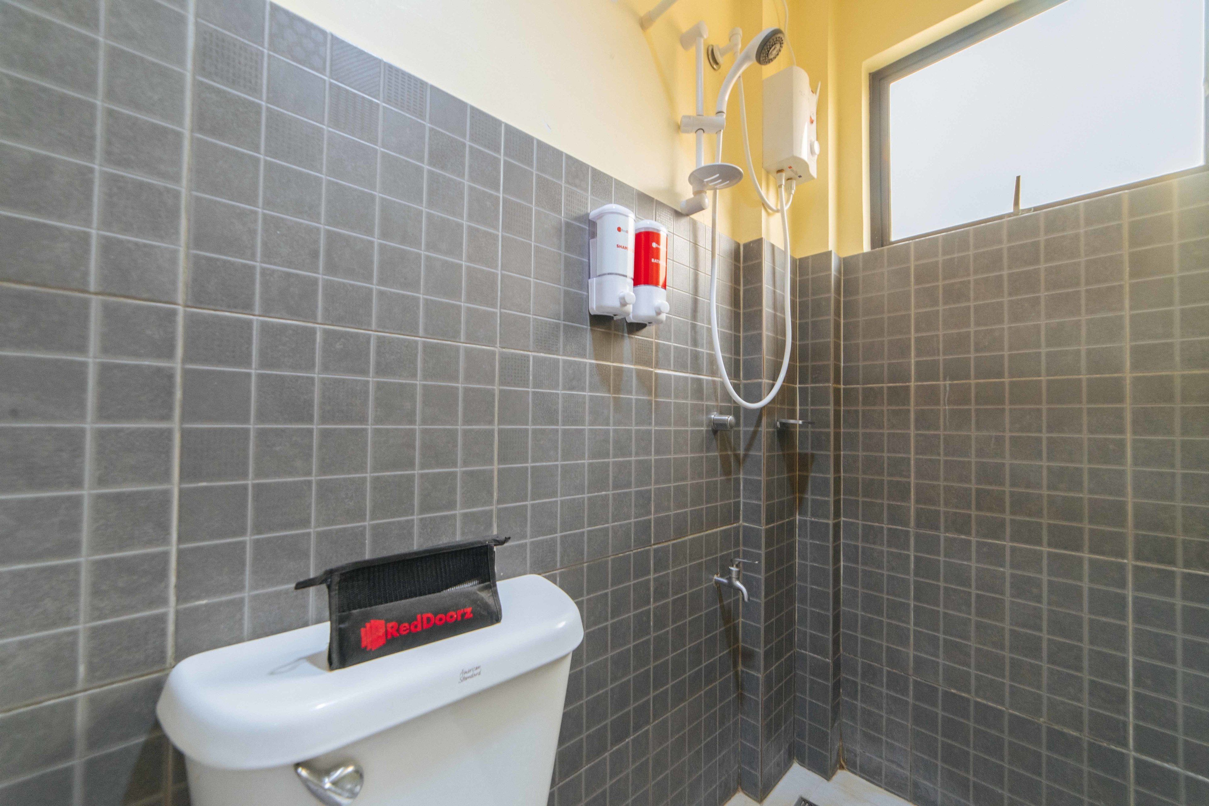 Un baño con paredes de azulejos grises, inodoro blanco, dispensador de jabón rojo y ducha.