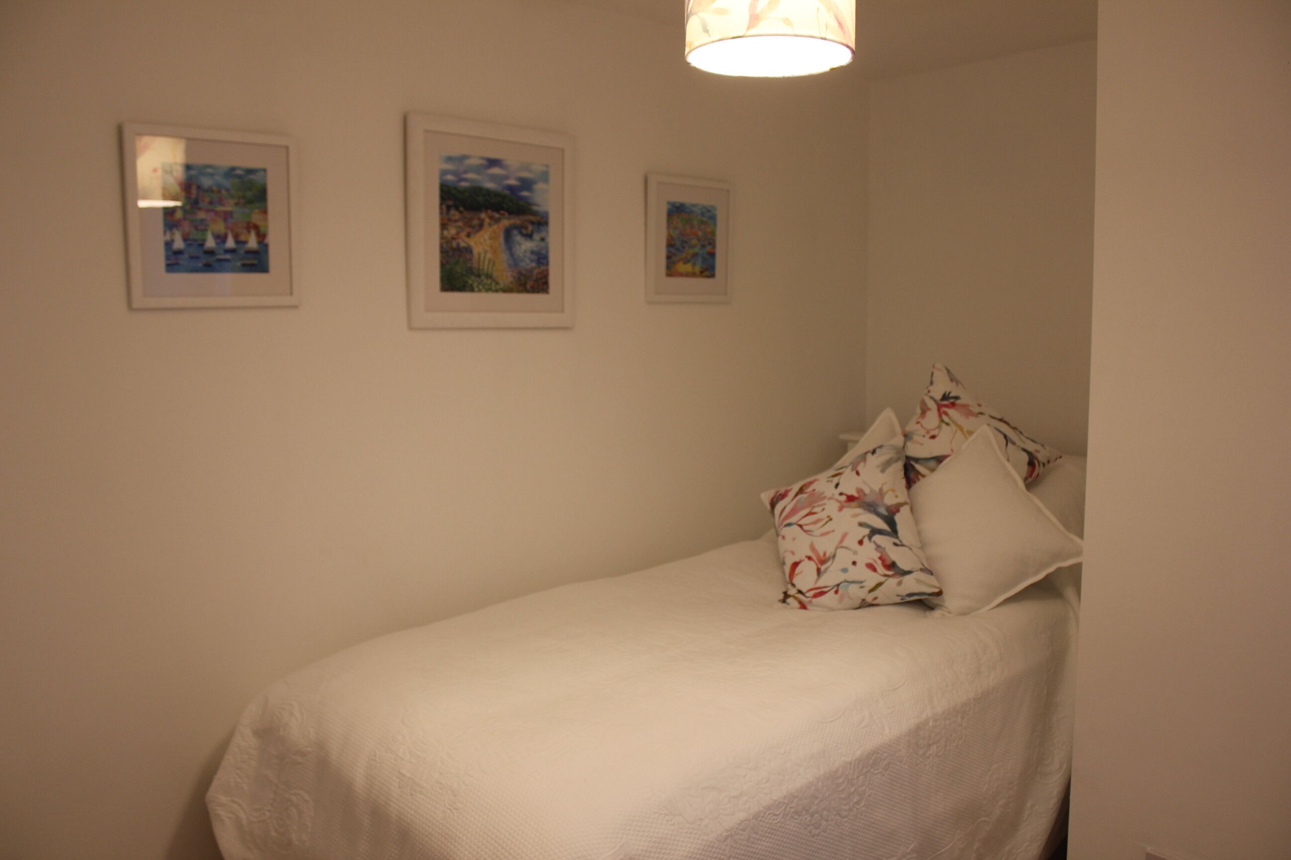 Un lit simple avec une couvre-lit blanche et deux oreillers, dont l’un est orné d’un motif floral. Trois œuvres d’art encadrées sont accrochées au mur au-dessus du lit.