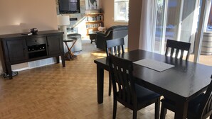 A dining area with a table and chairs, a TV mounted on a stone wall, a bookshelf, and a window with blinds. - Swimming pool, Family friendly home! (Penticton)