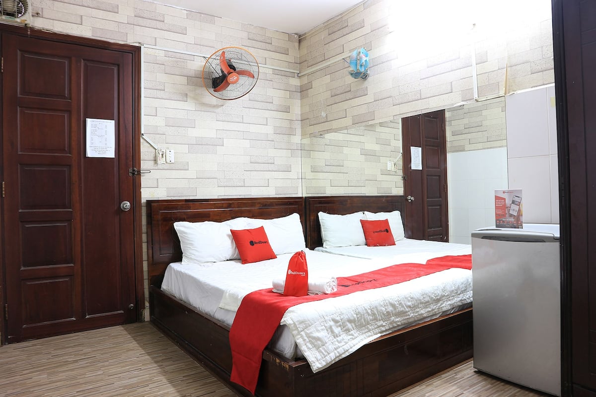 Ein Hotelzimmer mit zwei Betten, einem hölzernen Kopfteil, einer roten und weißen Bettdecke, einem kleinen Kühlschrank, einem Wandventilator und einer Wand mit Ziegeloptik.