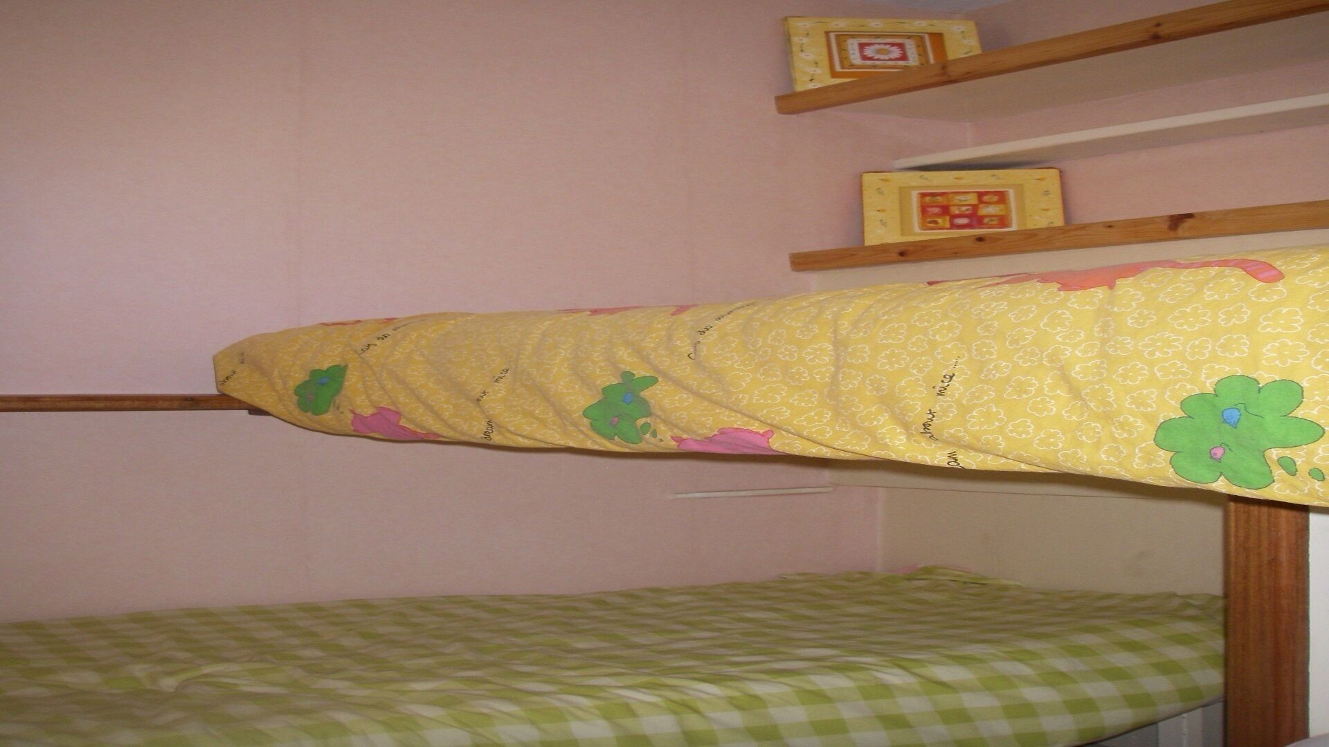 A bunk bed with a yellow mattress and a green checkered bottom. There are two shelves above the bed, one with a floral pattern and the other with a red and white design.