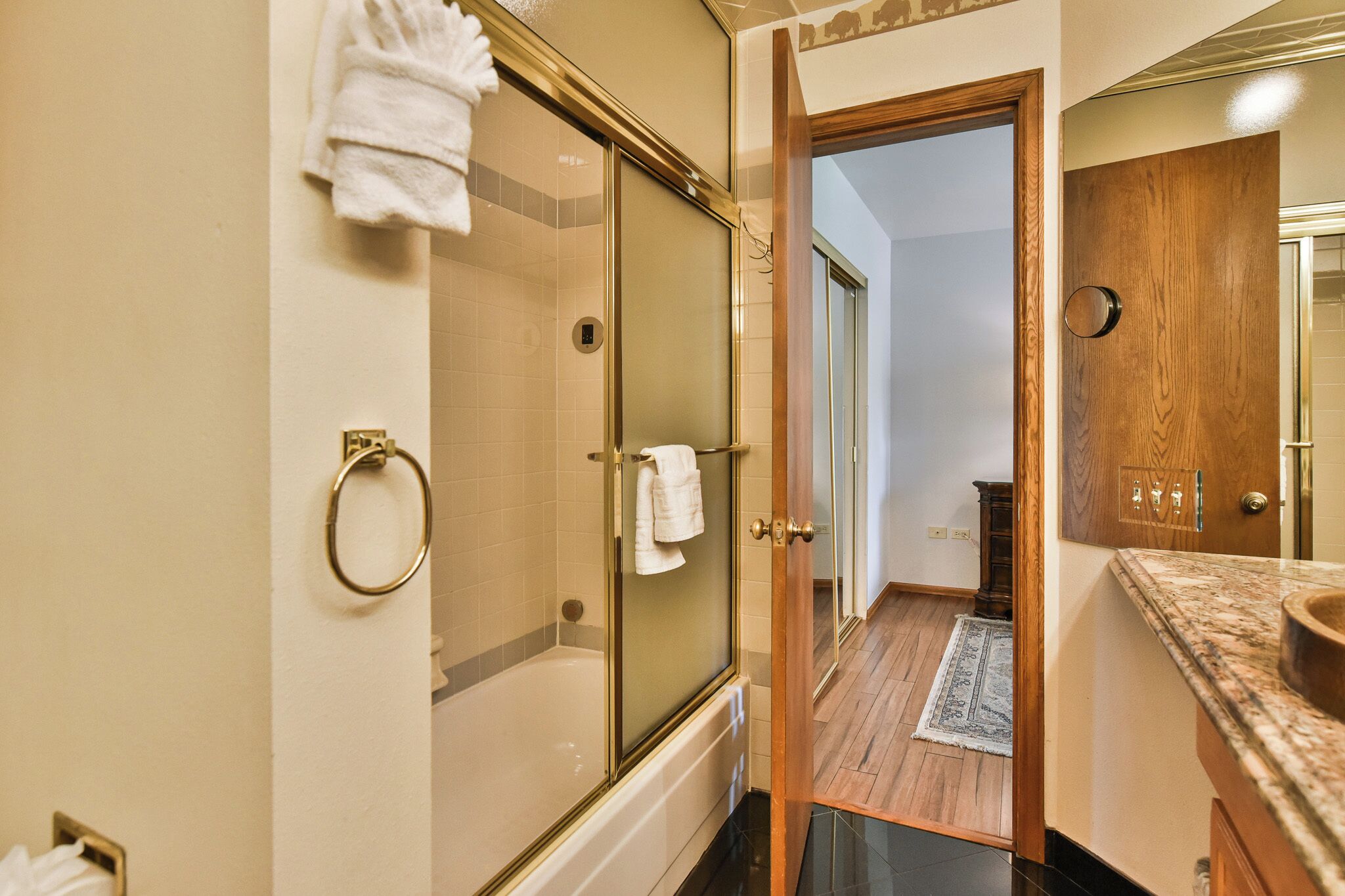 A bathroom with a glass-enclosed shower, a wooden vanity, and a visible hallway leading to another room.