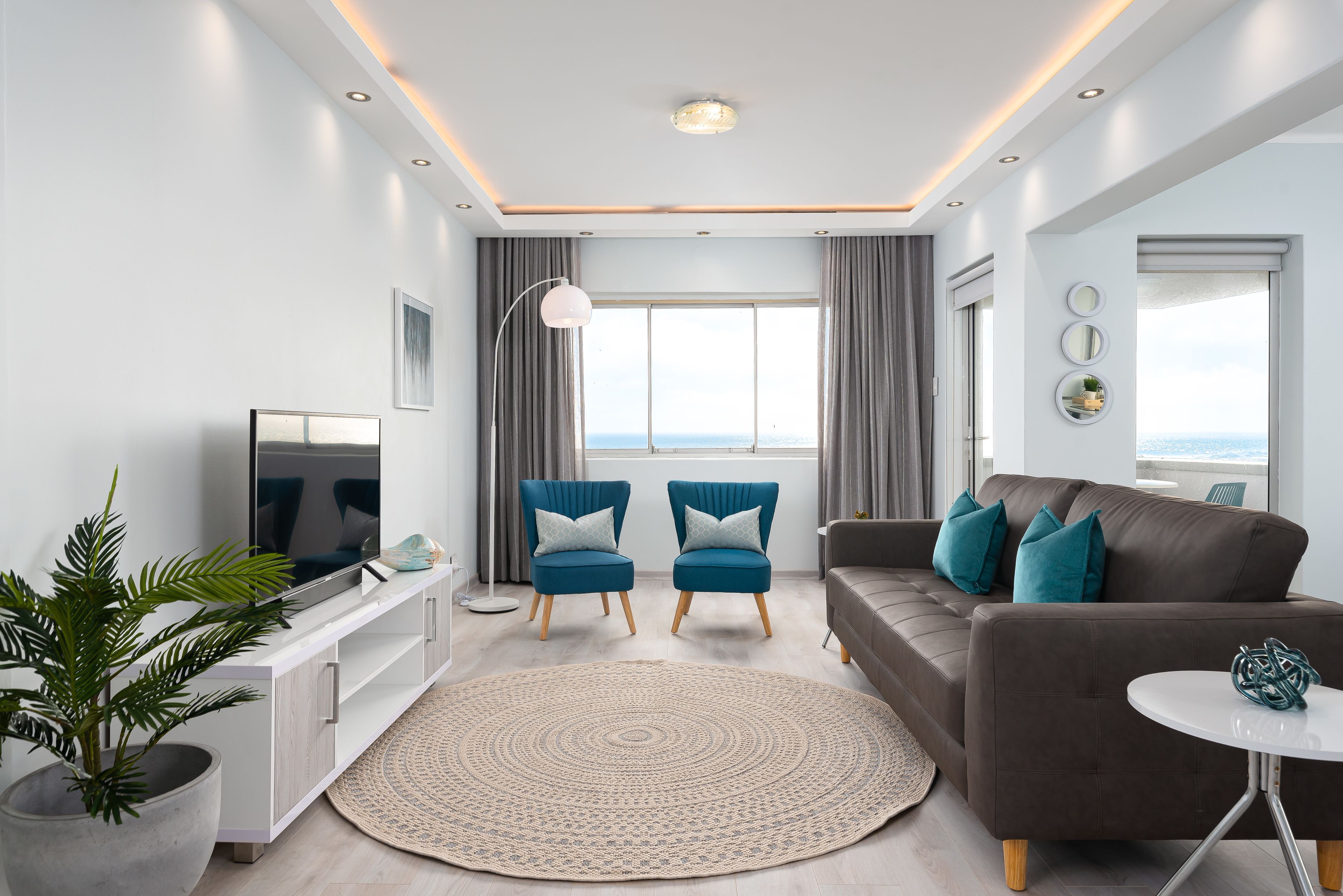 A modern living room with a TV, two blue armchairs, a brown sofa, a round rug, and a potted plant. - Saratoga 304 (Cape Town)