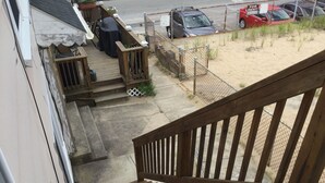 Un escalier en bois mène à une terrasse aménagée avec des sièges d’extérieur, d’où l’on peut admirer les voitures garées et la plage.
