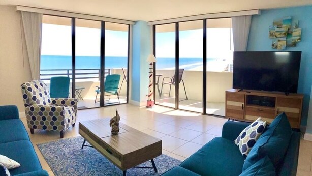 A living room with a sofa, chairs, a coffee table, and a TV. There is a balcony with a view of the beach. - OCEANFRONT NEAR THE PIER!OCEAN VIEW FROM EVERY ROOM! 2-2 WRAPAROUND BALCONY! (Daytona Beach)