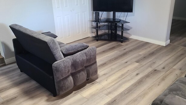 A living room with a grey recliner, a TV on a stand, and a wooden floor.