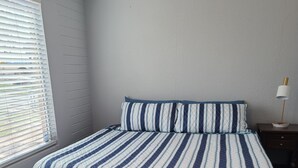 A bedroom with a bed, wooden flooring, a window with blinds, and a nightstand with a lamp.