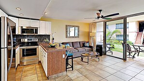 Cocina moderna con una isla central, zona de comedor con sofá y mesa, y puerta corrediza de vidrio que da a un patio exterior.