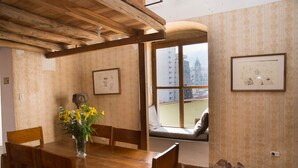 Een eetruimte met een houten tafel en stoelen, een vaas met bloemen, en een raam met uitzicht op een stadsgezicht.