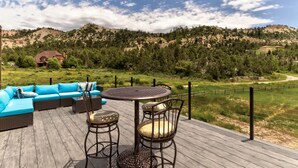 Um deck de madeira com móveis de exterior, incluindo um conjunto de sofás e uma mesa com cadeiras, com vista para uma paisagem montanhosa com árvores e uma casa ao fundo.