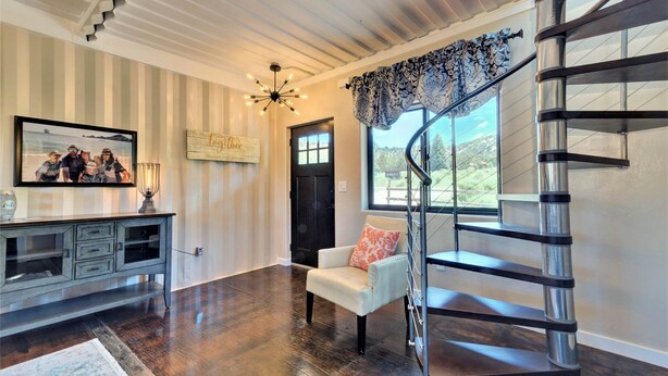 Uma sala de estar ampla com piso de madeira escura, uma poltrona branca com almofada estampada, uma mesa consola com objetos decorativos, uma fotografia emoldurada e uma escadaria em espiral.