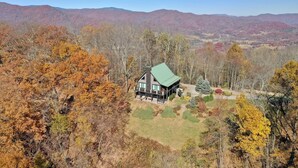 A cabin with a green roof surrounded by autumn foliage, set against a backdrop of mountains. - Don't Miss This View!!! Secluded Mountaintop Cabin- w/ Hot tub & Shuffleboard (Waynesville)