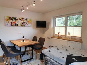 Um quarto com cama, janela com persianas, televisão na parede, mesa com cadeiras e um quadro na parede.