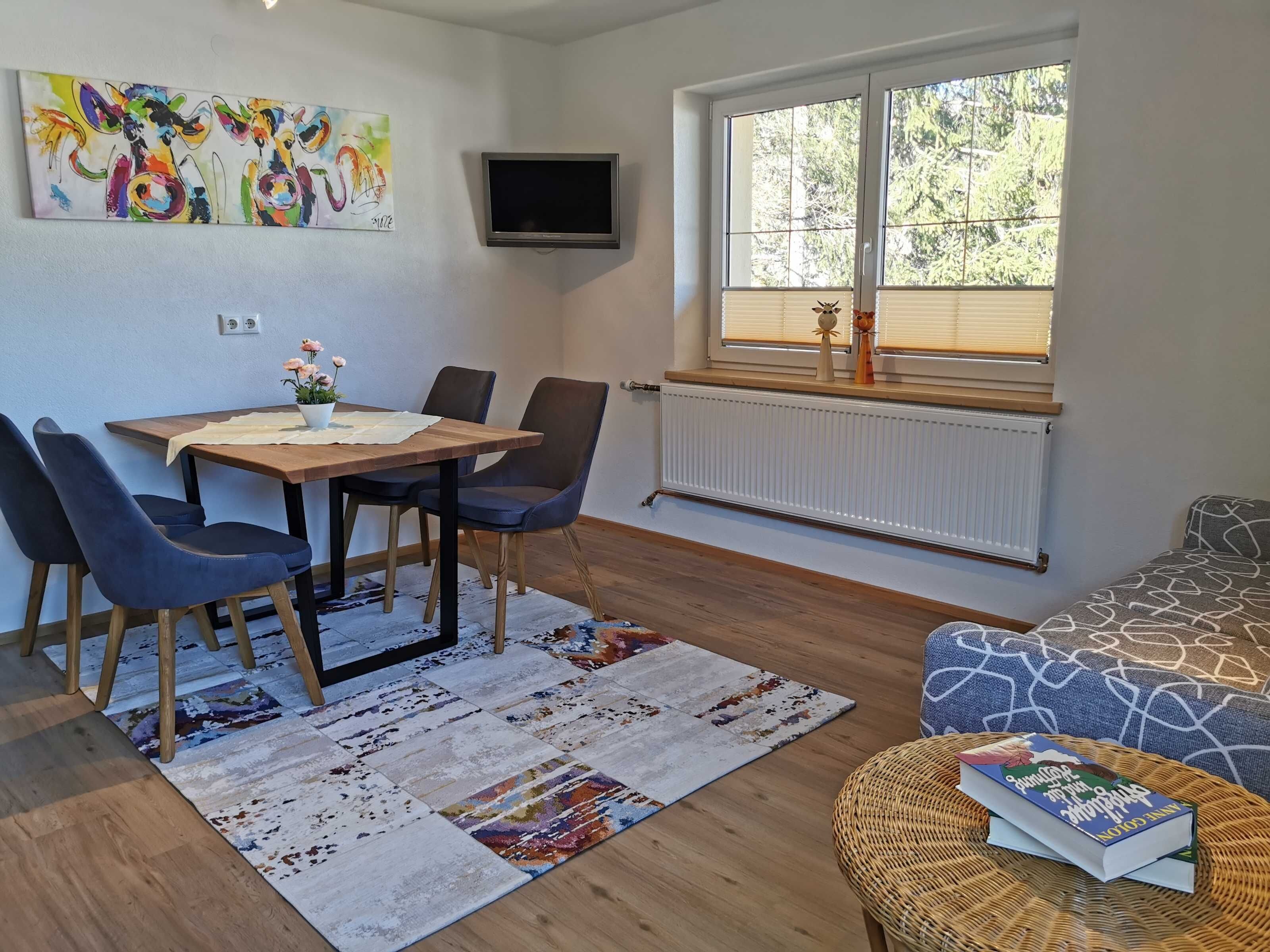 A dining area with a wooden table, blue chairs, a colorful rug, a TV mounted on the wall, and a window with blinds.