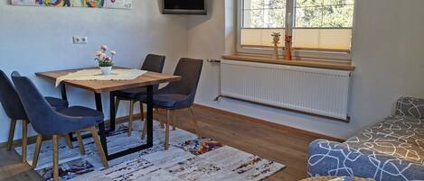 A dining area with a wooden table, blue chairs, a colorful rug, a TV mounted on the wall, and a window with blinds.