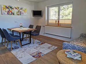 Ein Essbereich mit Tisch, Stühlen, einem Fernseher, einem Fenster mit Jalousien und einer Couch.