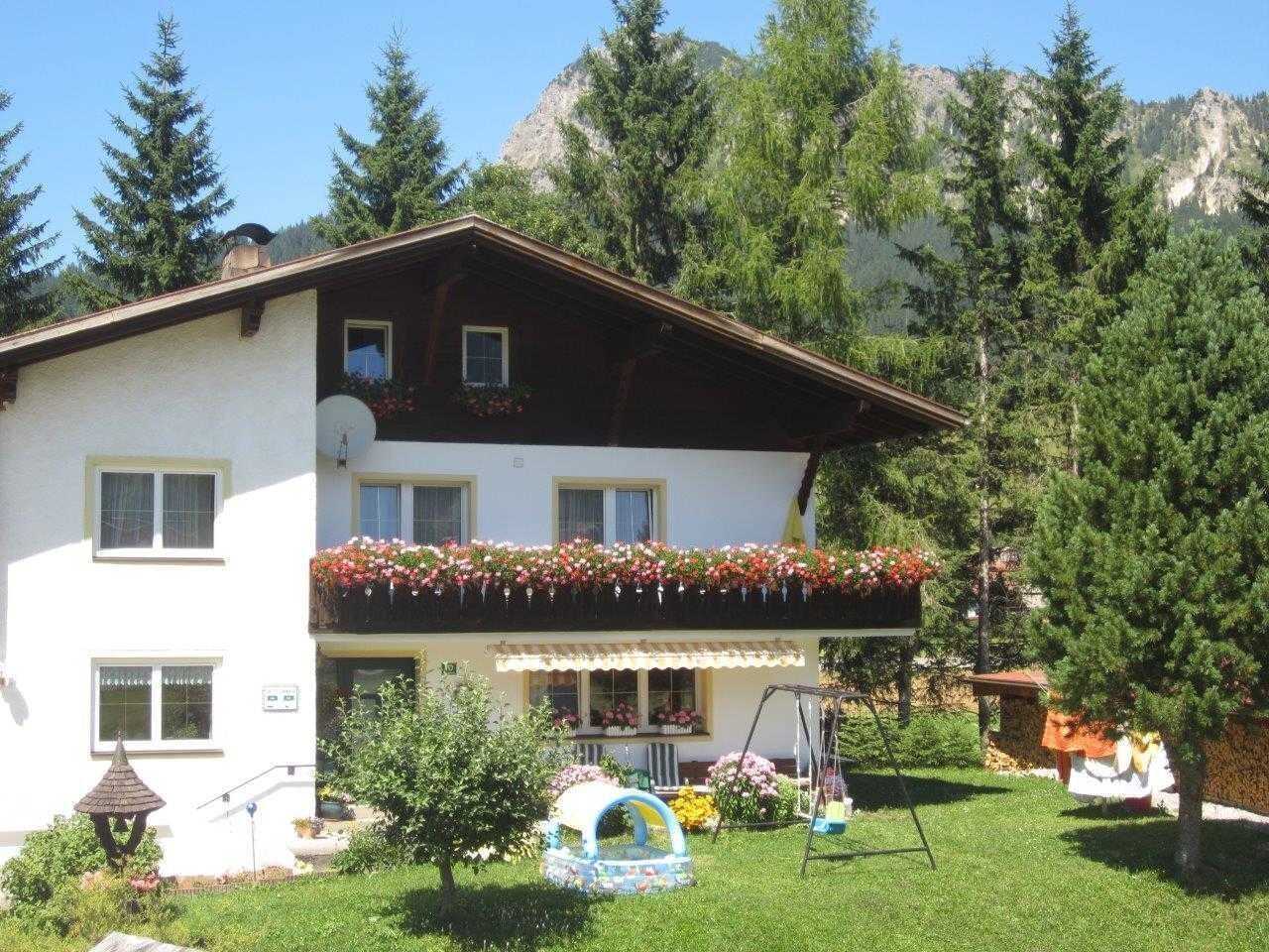Ein zweistöckiges Gebäude mit Balkon, umgeben von viel Grün und Bergen.