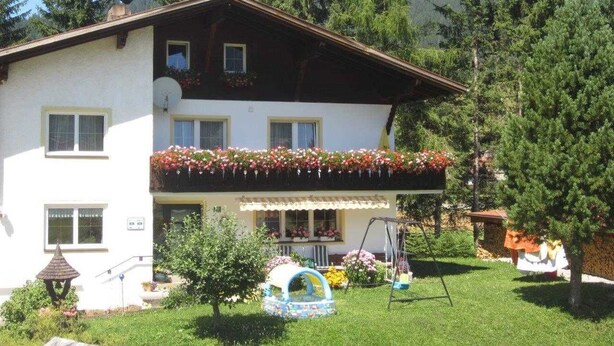 Ein zweistöckiges Gebäude mit Balkon, umgeben von viel Grün und Bergen.