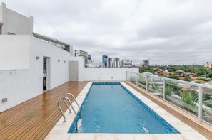 A rooftop swimming pool with a wooden deck, a glass balustrade, and a cityscape view in the background.