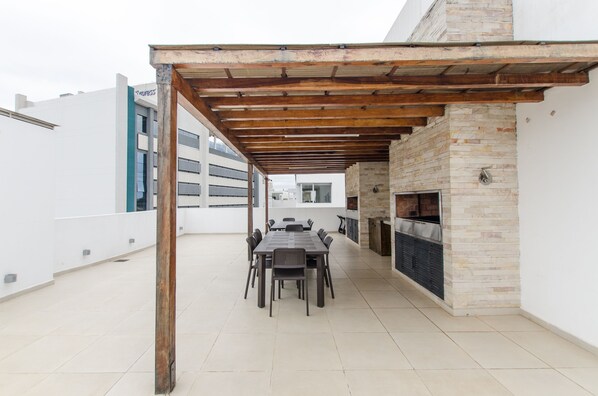 An outdoor patio with a long table and chairs, a brick fireplace, and a pergola structure.