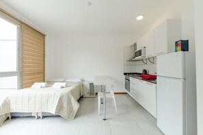 A compact room with a bed, a small table, a kitchenette, and a window with blinds.