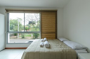 A hotel room with a large window, a bed with a beige quilt, and a wooden headboard. - Best location in Asuncion - Studio (Asunción)