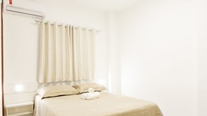 A neatly made bed with a beige bedspread and pillows, a small bedside table, and a window with curtains.