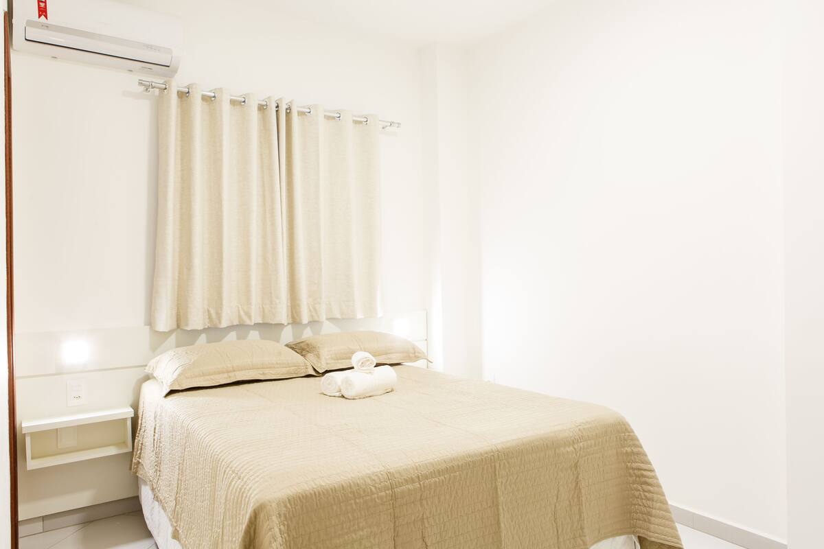 A neatly made bed with a beige bedspread and pillows, a small bedside table, and a window with curtains.