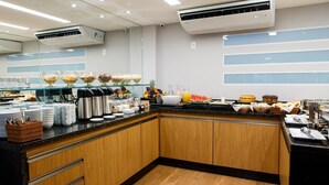 A modern hotel breakfast area with buffet-style food, coffee machines, and fruit displays.