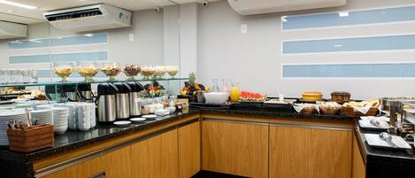 A modern hotel breakfast area with buffet-style food, coffee machines, and fruit displays.