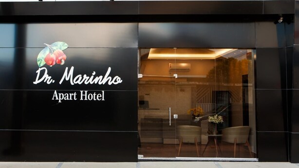 The exterior of Dr. Marinho Apart Hotel with a modern design, featuring a large glass entrance and a logo with fruit and leaves.
